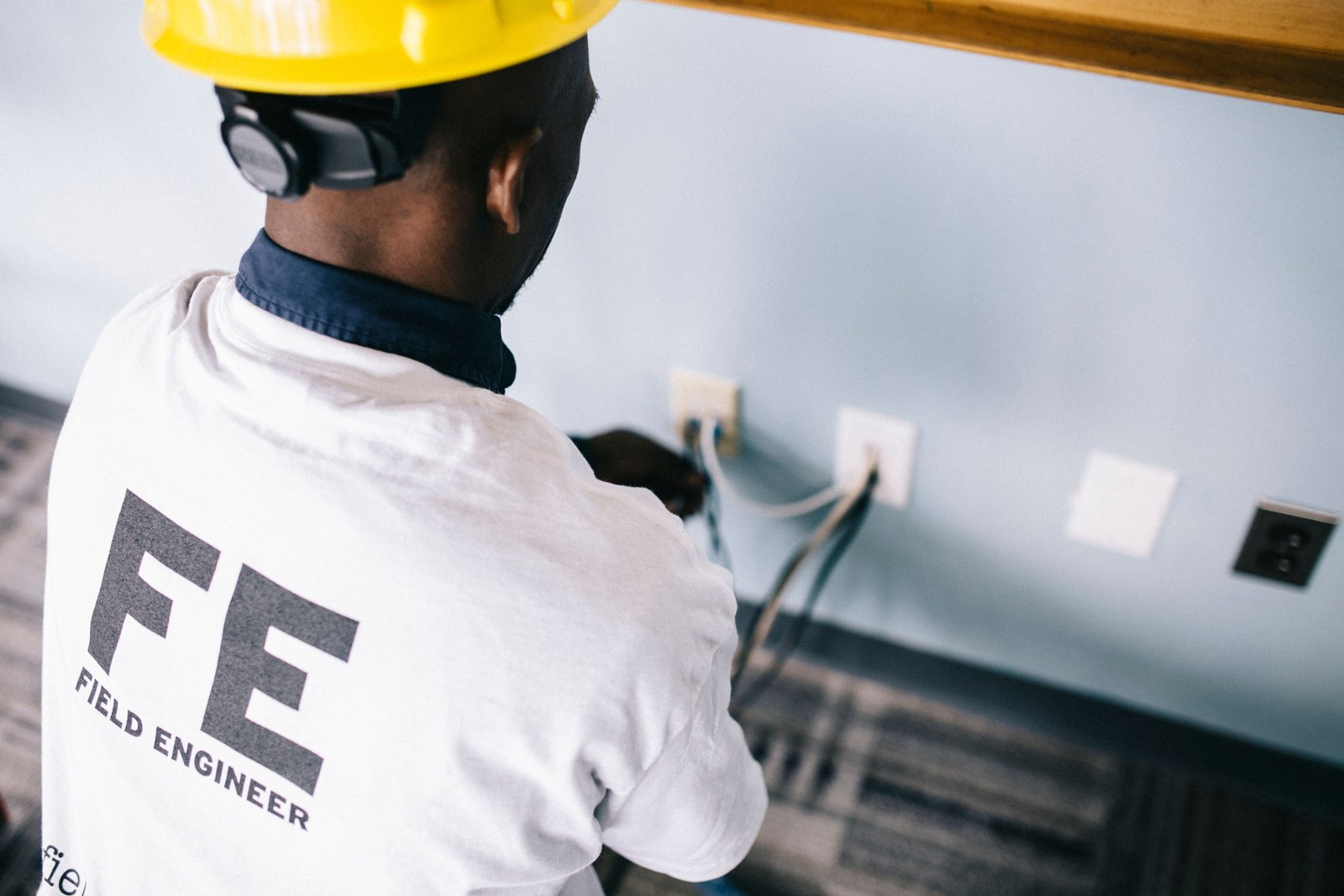 Electricista Homero Gonzalez Villar de Rena above back view unrecognizable black field engineer wearing white shirt and protective hardhat sitting on floor and checking voltage in wall plugs